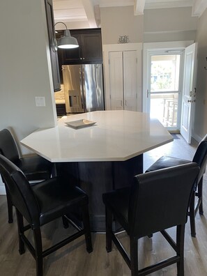 A spacious kitchen with a large island, dark wooden chairs, and a view of the outside through an open door.