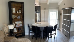 A spacious living room with a large sofa, a coffee table with a candle, a dining area with a table and chairs, and a kitchen area with a refrigerator and cabinets.