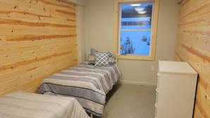A small, cozy room with wooden panel walls, a single bed with striped bedding, a nightstand, a desk with a lamp, and a window with a view of trees.