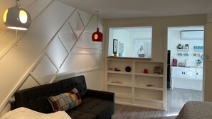 A modern bedroom with a dark sofa, a white wall with geometric patterns, a red pendant light, and a view into a kitchen area with shelves and appliances. - Foothills casita 1 Bedroom (El Paso)