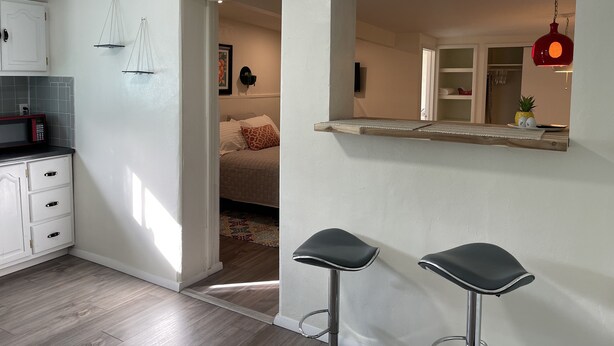A modern kitchen with white cabinets, a microwave, and a wooden countertop. There are two black bar stools and a hanging red pendant light. - Foothills casita 1 Bedroom (El Paso)