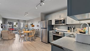 A modern kitchen with stainless steel appliances, a dining area with wooden furniture, and a view of the street outside. - A Wave From it All:  Condo on the Beach (Gulf Shores)