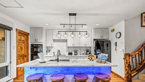 A modern kitchen with white cabinets, a marble island with bar stools, and a unique hexagonal pendant light. - Spacious 3B/3B Condo. Luxury Amenities. ShuffleBoard. HotTubs. 1st FLR. Location (Steamboat Springs)