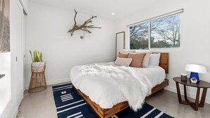 A modern bedroom with a large bed, a wooden headboard, a side table, a floor lamp, and a window with a view of trees. - Soul Shine| Amazing Views | Location | Remodeled, Soul Shine| Amazing Views | Location | Remodeled (Joshua Tree)