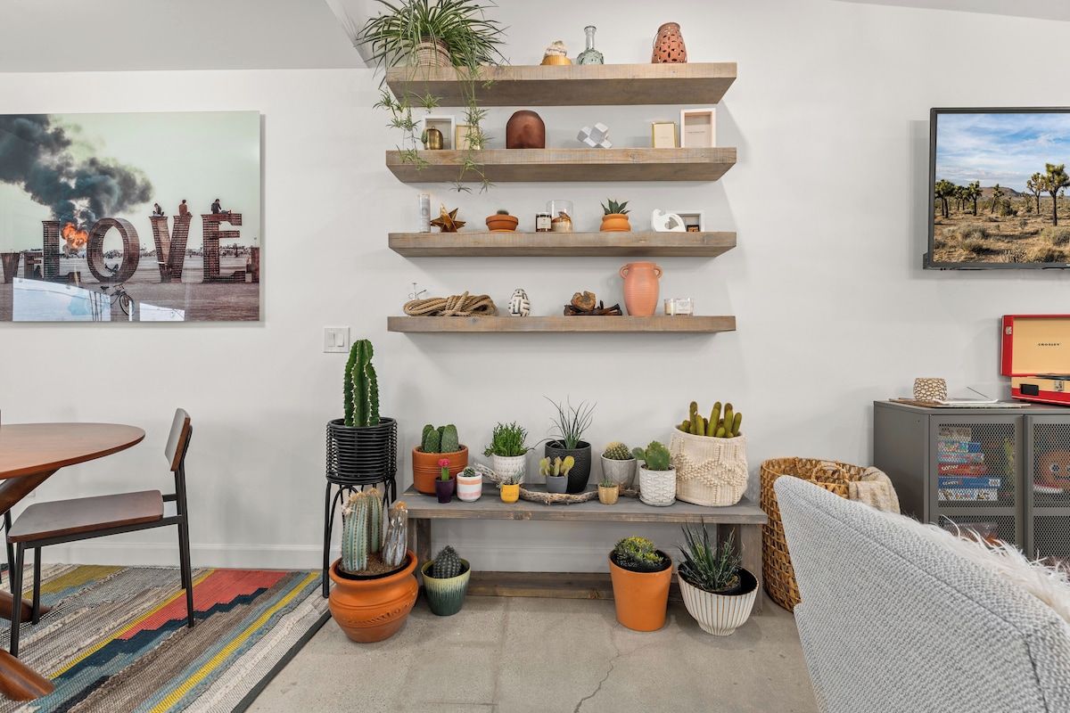 Un soggiorno con una mensola in legno che espone diverse piante e oggetti decorativi, una collezione di cactus e un televisore appeso al muro.