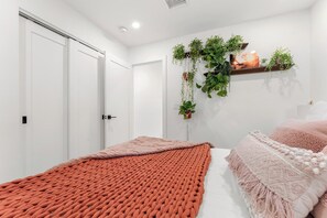Un dormitorio con cama, una colcha de punto rojo, un adorno macramé rosa y una estantería mural para plantas.
