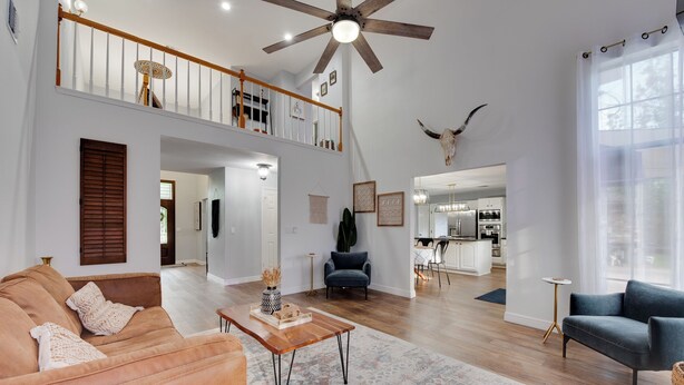 A spacious living room with a high ceiling fan, a wooden coffee table, and a large rug. A kitchen is visible in the background, and there is a balcony with a railing. - Family-Friendly 4BR w/ Heated Pool, Spa & Game Room near The Woodlands and IAH (Spring)