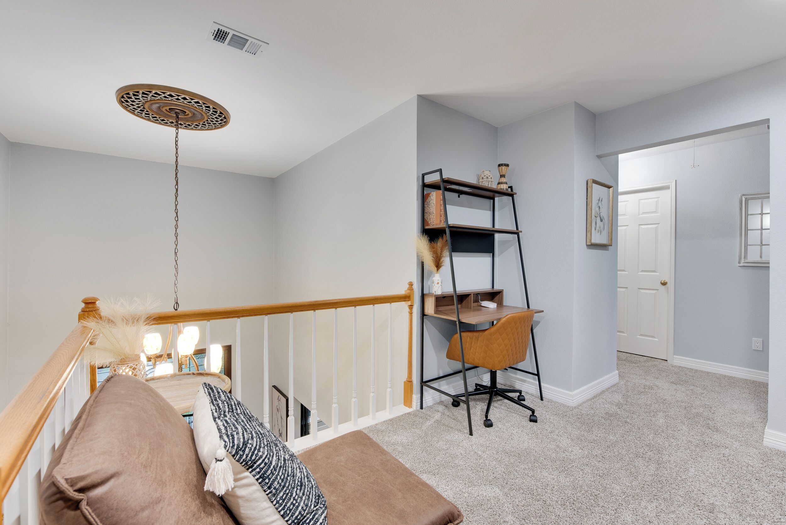 A staircase with a wooden handrail, a cushioned bench with pillows, a black metal desk with a chair, and a decorative ceiling light.