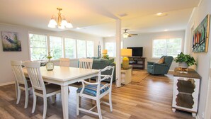 A spacious living area with a dining table, chairs, and a sofa. There are large windows allowing natural light, a ceiling fan, and a television.