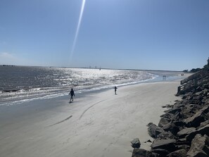 Ein Strand mit felsigem Ufer, blauem Himmel und einer entfernten Brücke.