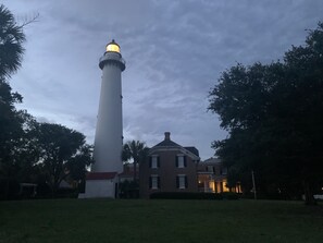Ein Leuchtturm mit Licht oben, ein Backsteinhaus im Vordergrund und Bäume ringsum.