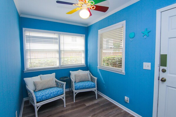 A room with two white wicker chairs with blue cushions, a ceiling fan, and two windows with white blinds.