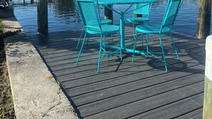 Eine Holzdar Terrasse mit türkisfarbenen Metallstühlen und einem Tisch, mit Blick auf ein ruhiges Gewässer, Bäume und eine entfernte Brücke.