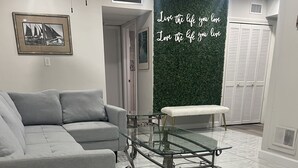 Ein modernes Wohnzimmer mit einem grauen Sofa, einem Glastisch und einer Bank. Eine grüne Wand mit einem motivierenden Zitat ist zu sehen.