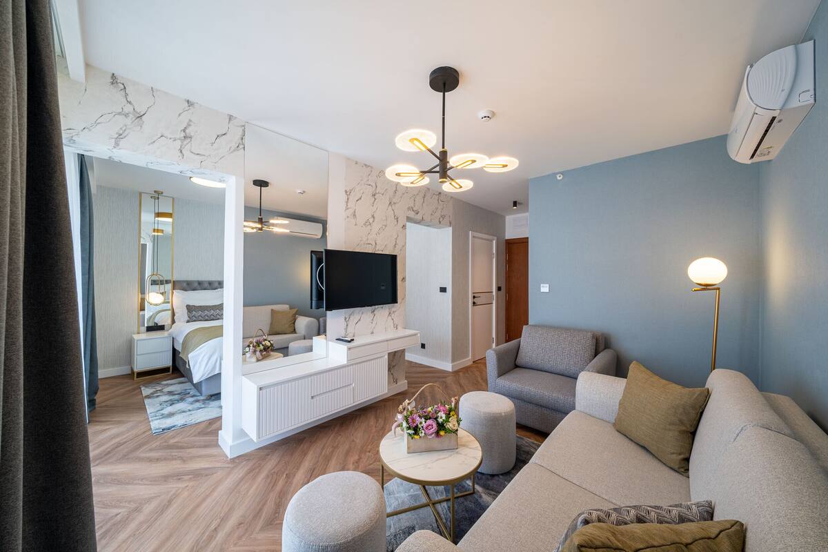 Una sala de estar moderna con sofá, reposapiés y una mesa de centro con flores. Hay un televisor de pantalla plana montado en la pared y una lámpara en el techo.
