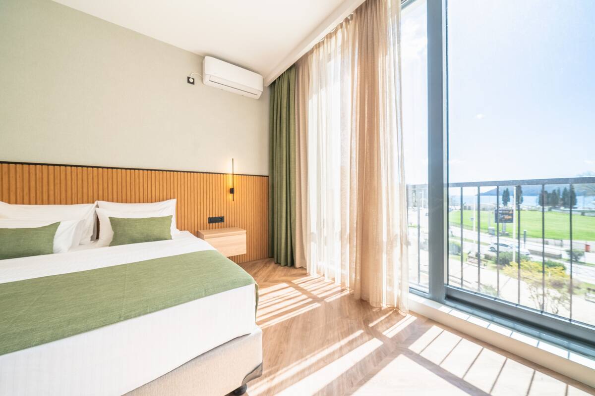 Habitación de hotel con una cama grande, una mesita de noche, aire acondicionado de pared y vistas a un paisaje urbano a través de un ventanal.