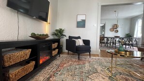 Een woonkamer met een open haard, een zwarte bank, een eethoek met een tafel en stoelen, en een aan de muur bevestigde televisie.