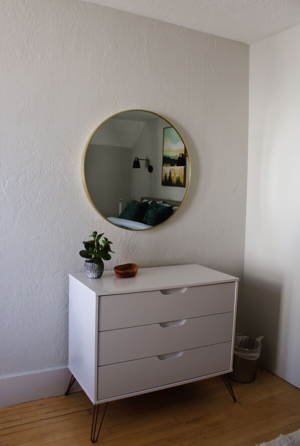 A white dresser with a round mirror, a small plant, and a bowl on top.