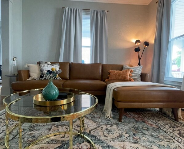 A living room with a brown leather sofa, a glass coffee table with a teal vase, and a patterned rug.