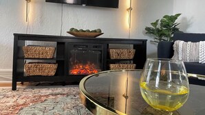 Een woonkamer met een open haard, een glazen salontafel met een wijnglas, een zwarte bank en een plant.