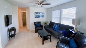 Un salon avec un ventilateur de plafond, un téléviseur, un canapé, une table basse et une lampe.