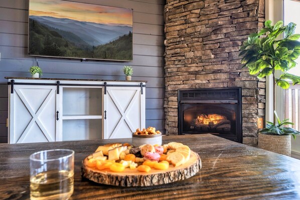 A rustic interior with a stone fireplace, a dining table with a fruit platter, and a mounted painting. - AMAZING Mountain Views Retreat | SemiSecluded/Games/FirePit/Upscale/HotTub (Sevierville)