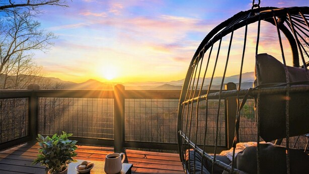 A wooden deck with a swing chair, a potted plant, and a mug on a table, overlooking a mountainous landscape during sunset.