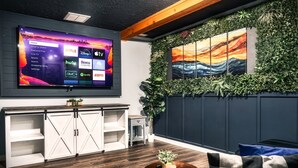 A modern living room with a large flat-screen TV displaying a streaming service interface, a wooden cabinet, a round coffee table with a candle, and a wall adorned with greenery and a painting.