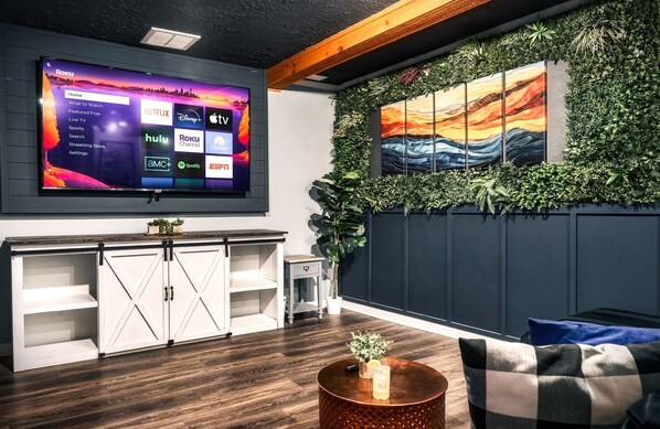 A modern living room with a large flat-screen TV displaying a streaming service interface, a wooden cabinet, a round coffee table with a candle, and a wall adorned with greenery and a painting. - AMAZING Mountain Views Retreat | Games/FirePit/Upscale/HotTub/SemiSecluded (Sevierville)