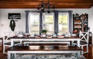 A dining area with a wooden table set for six, white chairs, a chandelier, a welcome sign, and a coffee bar with mugs.