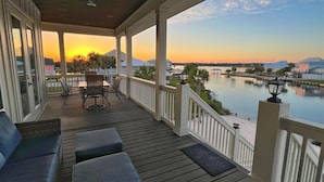 A covered deck with outdoor seating, a view of a calm body of water, and a sunset in the background. - The Shoreland Water-Front Home, Volleyball &Hottub (Gulf Shores)