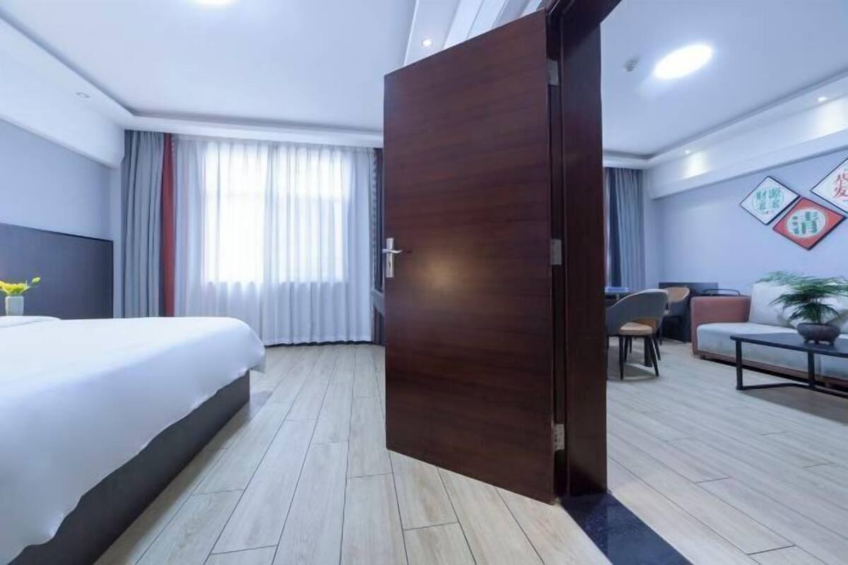 A hotel room with a bed, a wooden door, a seating area with a sofa and chairs, and a window with curtains.