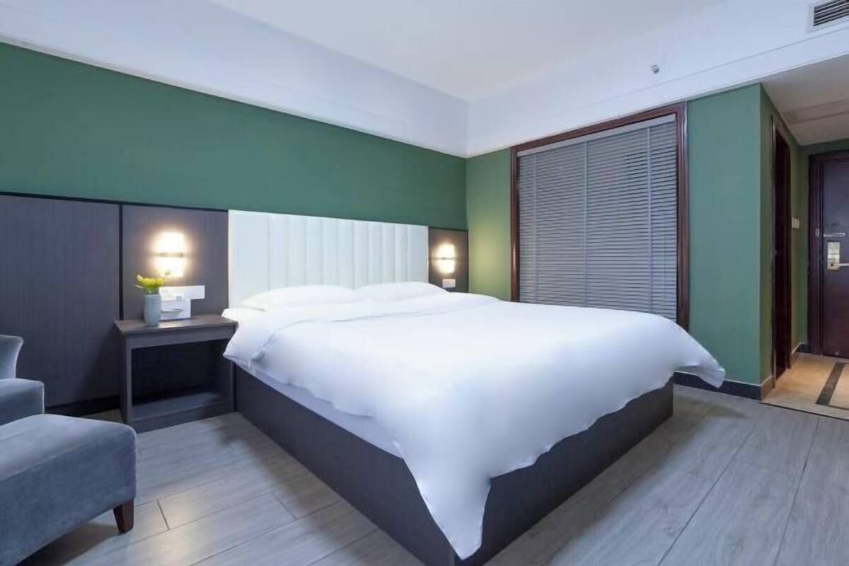 A modern hotel room with a large bed, bedside tables, a chair, and a window with blinds.