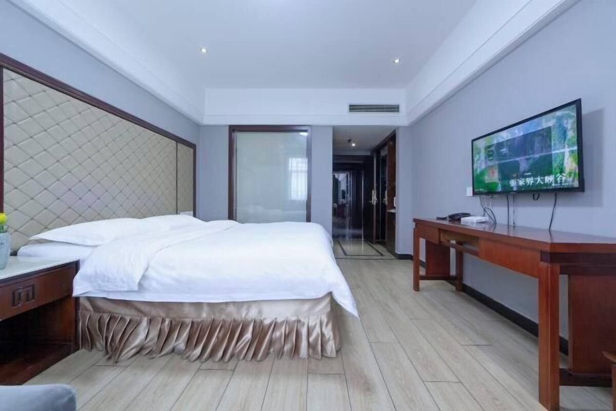 A hotel room with a bed, a TV mounted on the wall, a wooden desk, and a mirror.