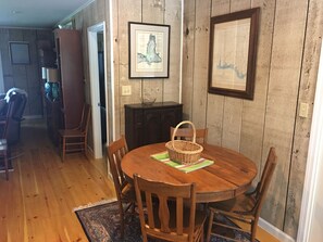 A dining area with a round wooden table, four chairs, a wicker basket, and two framed maps on the wall. - Beautiful , private Bon Secour riverfront home with spacious screened porch (Bon Secour)