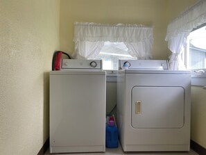A laundry area with two washing machines, a window with white curtains, and a blue detergent bottle.