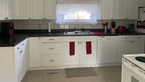 A kitchen with white cabinets, a black countertop, a window with a curtain, and a red towel hanging on the door. - Perfect Family Vacation House - Walk to Beach (Sylvan Beach)