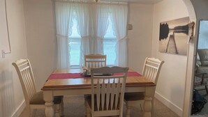A dining area with a wooden table and four chairs, a window with sheer curtains, a ceiling light, and a painting on the wall. - Perfect Family Vacation House - Walk to Beach (Sylvan Beach)