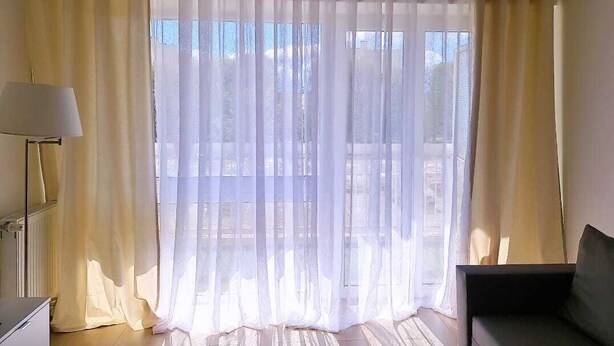 Ein Zimmer mit einem großen Fenster, das mit transparenten Vorhängen drapiert ist, einer Stehlampe und einem Sofa.
