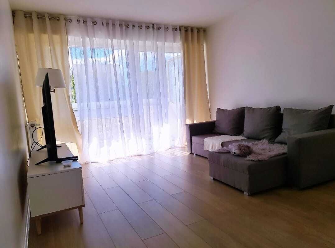 Ein Wohnzimmer mit einer Couch, einem Fernseher auf einem Schrank und einem Fenster mit durchsichtigen Vorhängen.