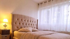 Ein Schlafzimmer mit einer gesteppten Kopfteil, einem Nachttisch mit Lampe und einem Fenster mit durchsichtigen Vorhängen.