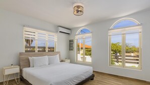 A bedroom with a large bed, two bedside tables, an air conditioning unit, and two windows with arched tops.