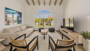 A spacious living room with a large arched window, a flat-screen TV, a beige sofa, two wicker chairs, and a coffee table.