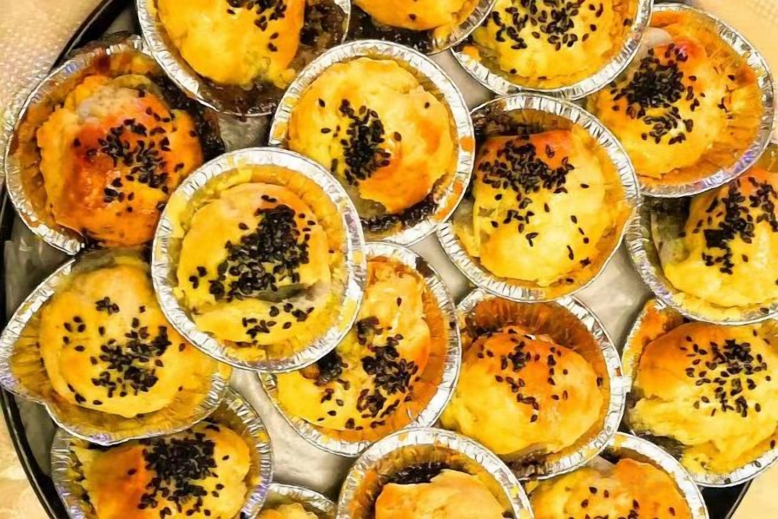 A tray of mini pies with a golden-brown crust, some topped with black seeds.