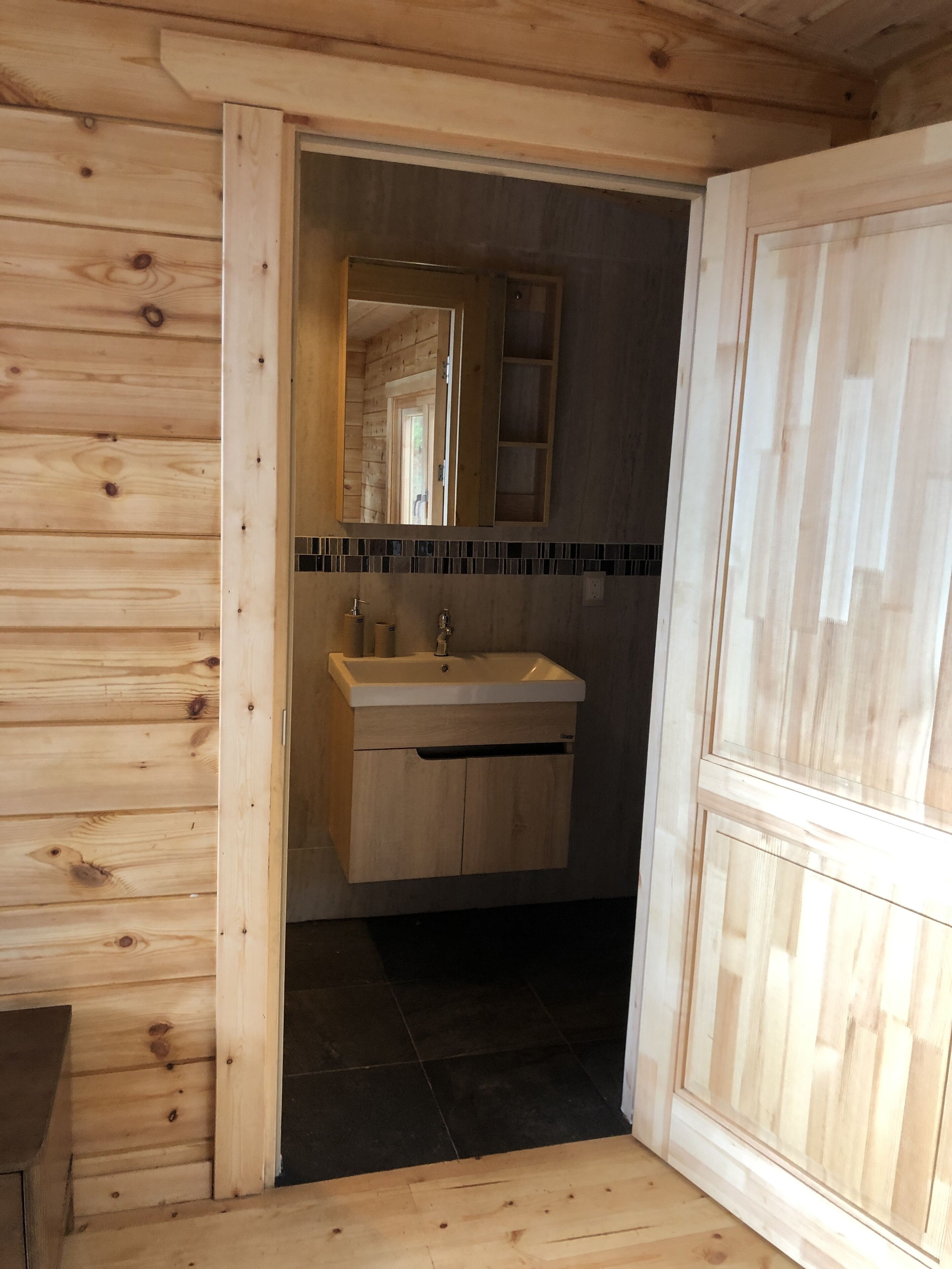 Interior de una cabaña de madera con un baño que incluye un lavamanos, espejo y suelo de baldosas.