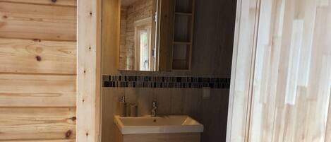 Interior de una cabaña de madera con un baño que incluye un lavamanos, espejo y suelo de baldosas.
