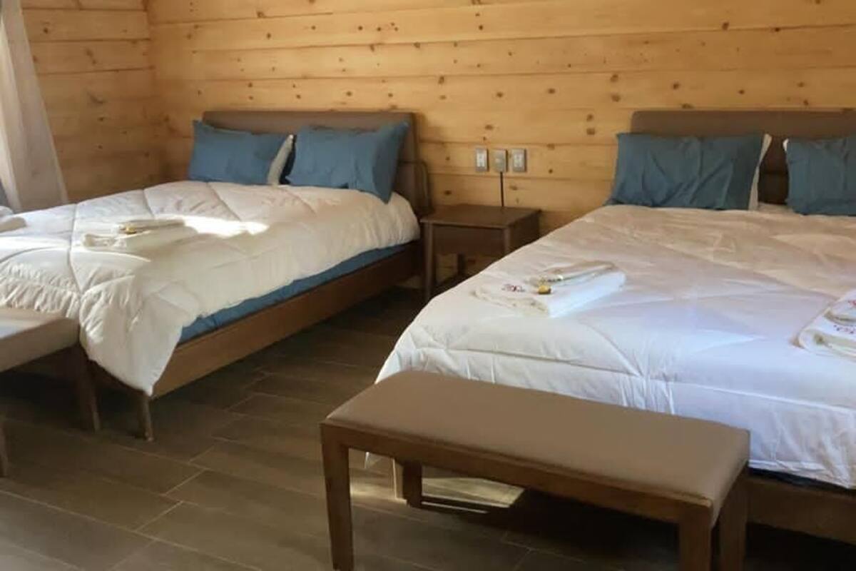Quarto com duas camas de solteiro e uma cama grande, cada uma com lençóis brancos e almofadas azuis. Há um banquinho de madeira no centro.