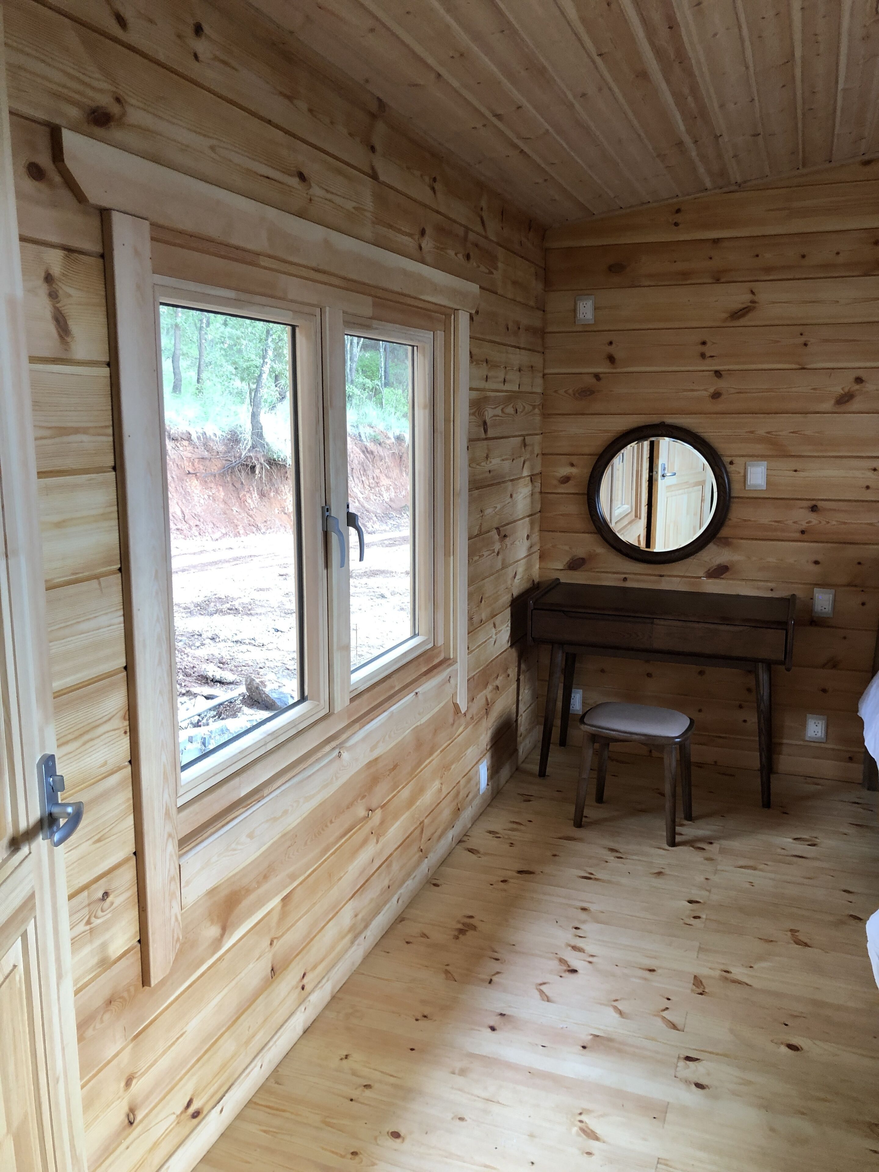 A wooden cabin interior with two large windows, a round mirror, and a small desk with a chair.