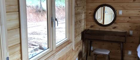 A wooden cabin interior with two large windows, a round mirror, and a small desk with a chair.
