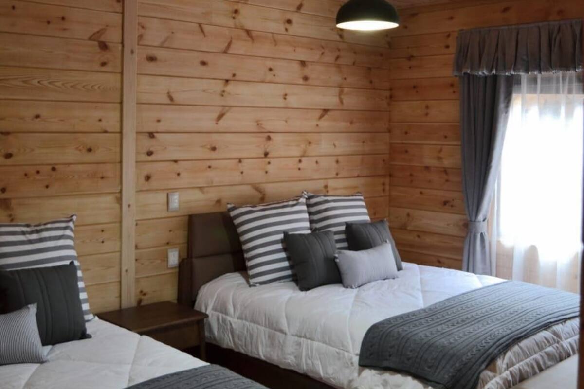 Quarto estilo cabana com duas camas, paredes de madeira, luminária pendente e janela com cortinas.